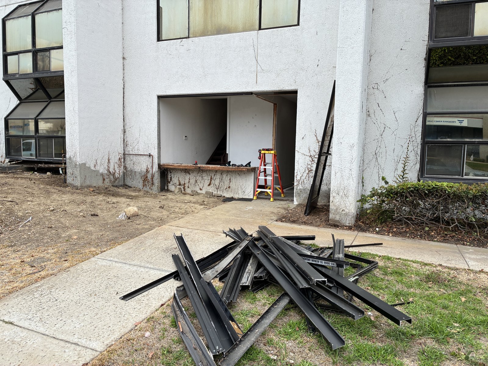 The original aluminum storefront glazing removed from the building and stacked on the grass, with a wall opening cut for a new entrance.